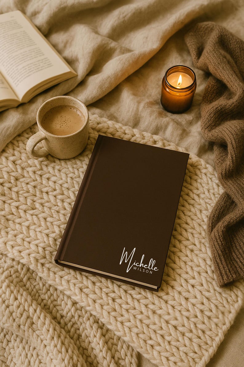 May include: A brown journal with the name "Michelle Wilson" in white script is displayed on a cream-colored knitted blanket. A cup of coffee, a lit candle, and an open book are also visible, creating a cozy scene.