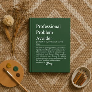 May include: A green hardcover book titled "Professional Problem Avoider" with a definition and the name "Stacy" below. The book is on a textured surface with art supplies and dried plants.