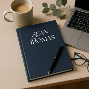 Peut inclure: Un journal bleu marine avec le nom "SEAN THOMAS" en lettres blanches. Un stylo noir est pos&eacute; sur le journal, &agrave; c&ocirc;t&eacute; d'un ordinateur portable, d'une tasse de caf&eacute; et de lunettes. Le journal est sur une surface en bois clair.