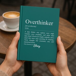 May include: A teal-colored book titled "Overthinker" with a definition of the word. The book is held in someone's hands, with a cup of coffee in the background. The definition includes the phrase "Also known as: Stacy."