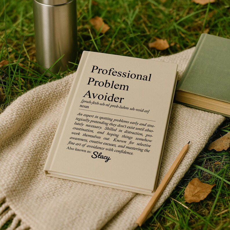 May include: A book titled "Professional Problem Avoider" with a definition, lying on a beige blanket. A stainless steel tumbler and a green book are in the background. A pencil rests on the blanket.