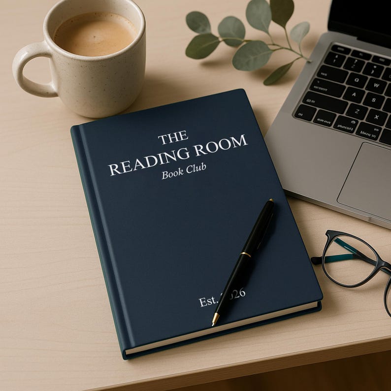 May include: A navy blue book with the text "THE READING ROOM Book Club" in white, and "Est. 26". A black pen lies on the book, beside a laptop, a coffee cup, and spectacles.