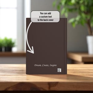 May include: A brown book with the text "Dream, Create, Inspire" on the cover. The image includes the text "You can add a custom text to the back cover" with an arrow pointing to the book. The book is on a wooden surface.