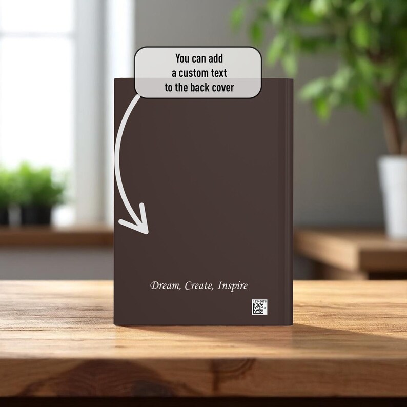 May include: A brown book with the text "Dream, Create, Inspire" on the cover. The image includes the text "You can add a custom text to the back cover" with an arrow pointing to the book. The book is on a wooden surface.