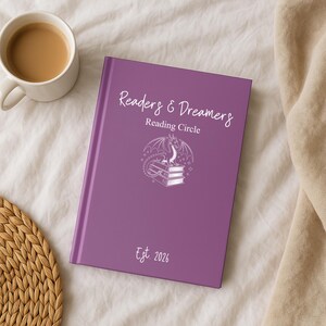 May include: A purple journal with the text "Readers & Dreamers Reading Circle" and a dragon illustration. The journal also has the text "Est. 2026". A white mug with a brown liquid sits to the left of the journal.