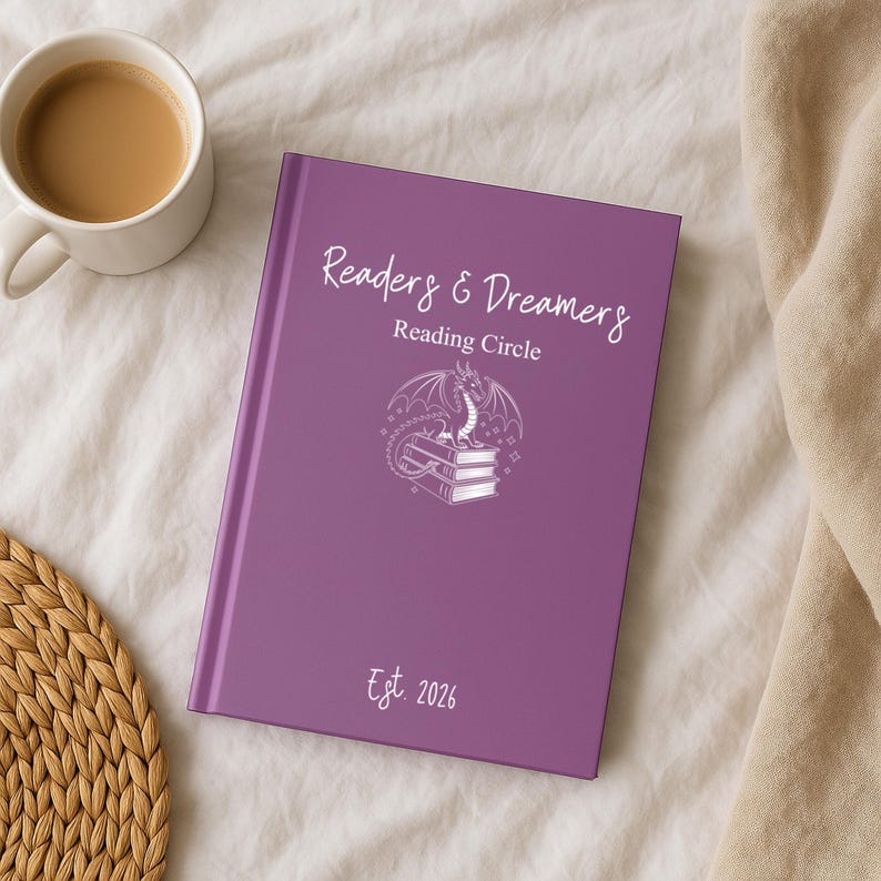 May include: A purple journal with the text "Readers & Dreamers Reading Circle" and a dragon illustration. The journal also has the text "Est. 2026". A white mug with a brown liquid sits to the left of the journal.