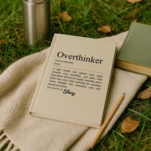 May include: A beige book titled "Overthinker" with a definition, resting on a beige blanket. A stainless steel tumbler and a green book are also visible. A pencil lies nearby on the grass.
