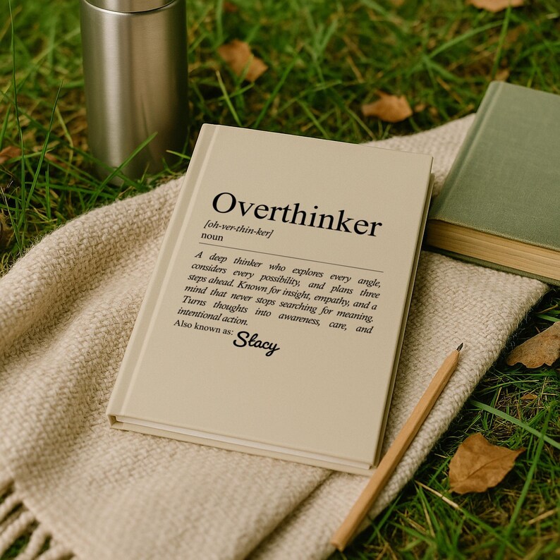 May include: A beige book titled "Overthinker" with a definition, resting on a beige blanket. A stainless steel tumbler and a green book are also visible. A pencil lies nearby on the grass.