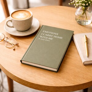 May include: A hardcover book with the title "EMOTIONS I CARRY HOME WITH ME" by Emily Wilson, in white text on a sage green cover. The book sits on a wooden table next to a cup of coffee, a saucer, and a pair of glasses.