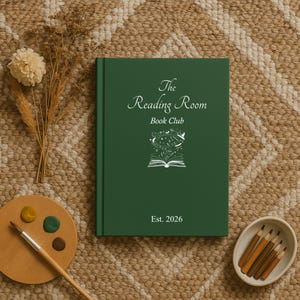 May include: A dark green book with the text "The Reading Room Book Club" and "Est. 2026" in white. The cover features a white illustration of an open book with celestial elements. The book is on a textured, neutral-toned surface.
