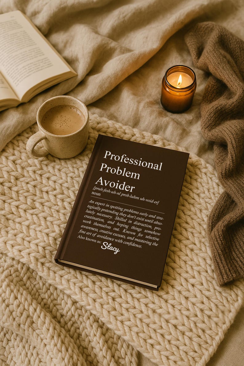 May include: A brown book titled "Professional Problem Avoider" with text and a definition, next to a cup of coffee, a lit candle, and a cozy blanket. The book is also known as "Stacy".