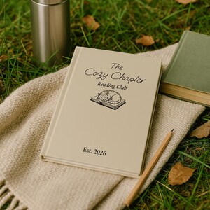 May include: A beige book titled "The Cozy Chapter Reading Club" with a sleeping cat illustration, a pencil, and a stainless steel tumbler on a woven blanket. A green book is visible in the background. The book is dated "Est. 2026".