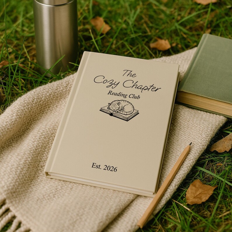 May include: A beige book titled "The Cozy Chapter Reading Club" with a sleeping cat illustration, a pencil, and a stainless steel tumbler on a woven blanket. A green book is visible in the background. The book is dated "Est. 2026".