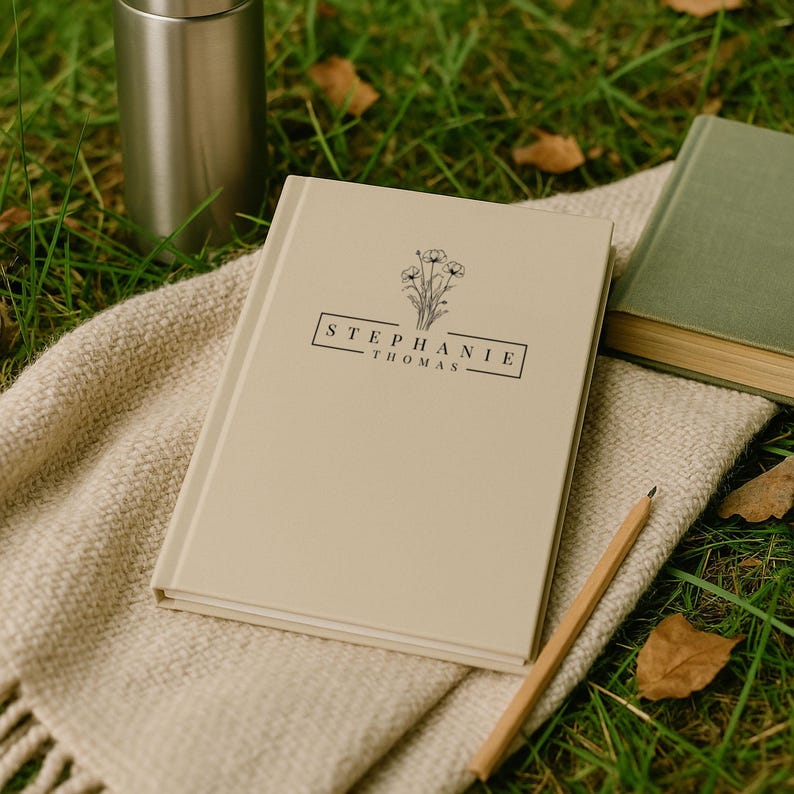 Peut inclure: Un journal beige avec le nom "STEPHANIE THOMAS" imprim&eacute; sur la couverture, accompagn&eacute; d'une illustration florale. Le journal est pos&eacute; sur une couverture tiss&eacute;e beige, &agrave; c&ocirc;t&eacute; d'un crayon, d'un livre vert et d'un gobelet argent&eacute;.