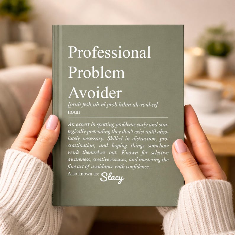 May include: A sage green book titled "Professional Problem Avoider" is held by a person. The book's definition includes the phrase "Skilled in distraction, procrastination." The author is also known as Stacy.