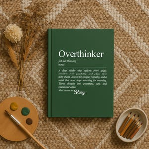 May include: A green hardcover notebook with the word "Overthinker" printed on the cover. The definition of "Overthinker" is printed below the title. A wooden palette with paint and pencils are next to the notebook.
