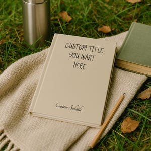 May include: A beige notebook with the text "CUSTOM TITLE YOU WANT HERE" and "Custom Subtitle" sits on a cream-colored blanket. A pencil and a green book are also present. A silver thermos is in the background.