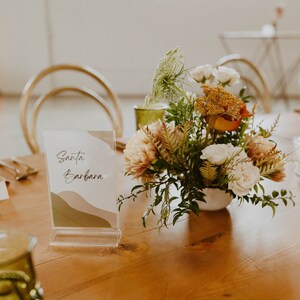 May include: A table setting with a clear acrylic sign that reads "Santa Barbara" and a floral centrepiece with white, orange, and green flowers in a white ceramic vase.