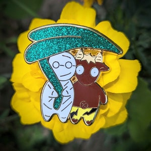 Könnte beinhalten: Eine goldene Emaille-Anstecknadel mit einer weißen Mumie mit einem grünen Glitzer-Regenschirm und einem braunen Monster mit Hörnern und gelben Blitzen.