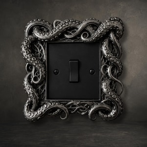 Peut inclure: Une plaque d'interrupteur carrée dotée d'un cadre argenté détaillé sculpté avec des tentacules de pieuvre entrelacés. La plaque centrale est noir mat avec un interrupteur à bascule vertical unique et deux vis de fixation noires visibles.