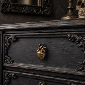 May include: A dark charcoal black wooden chest of drawers featuring an ornate, raised, antique gold-toned anatomical heart-shaped knob. The drawer front displays intricate carved scrollwork detailing at the corners and a textured wood grain finish.