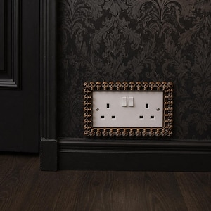 May include: A rectangular double electrical wall socket cover plate featuring a decorative border of small, metallic bronze-coloured skulls. The central white plastic socket plate includes two standard three-pin outlets and two rocker switches.