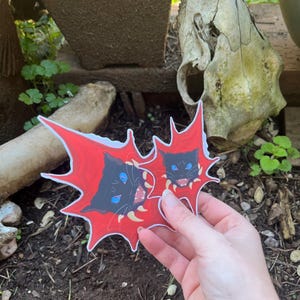 May include: Two stickers featuring black cats with blue eyes and fangs, set against a red burst design. The stickers are held in front of a natural outdoor background.