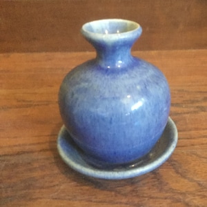 May include: A small blue ceramic vase with a round body and a narrow neck. The vase sits on a matching blue saucer.