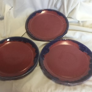 May include: Three ceramic plates with a deep red center and a dark blue, textured rim. The plates are round and have a simple, elegant design. The plates are arranged on a white surface.