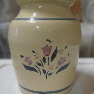 May include: A cream-colored ceramic crock with a speckled finish, featuring a hand-painted floral motif of pink tulips and dark blue leaves. The vessel has a wide mouth, a tapered body, and two horizontal dark blue bands encircling the top and bottom.