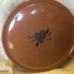 May include: A round, glazed ceramic plate with a speckled, earthy brown finish. The center of the plate features a hand-painted, dark brown heart icon with small, stylized wing-like markings on either side, creating a simple, centered design.