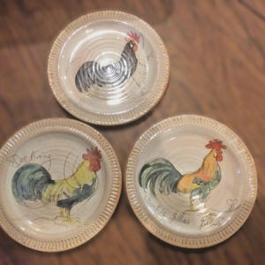 May include: Three decorative ceramic plates with rooster illustrations. Each plate features a cream-colored base with a detailed rooster design in shades of black, blue, yellow, and red. The plates have a textured rim.