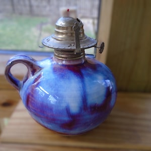 May include: A small, round oil lamp with a handle, featuring a blue and purple glazed ceramic body. The lamp has a silver-colored metal top with a wick and a small knob for adjusting the flame. The lamp is sitting on a wooden surface.