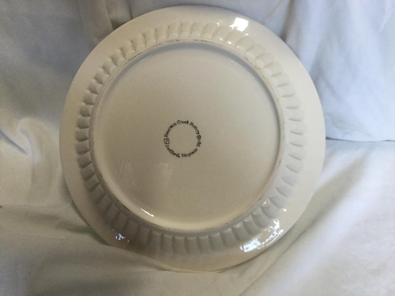 May include: White ceramic plate with a scalloped edge. The bottom of the plate has the text "James Creek Pottery, Stafford, Virginia" printed in black.
