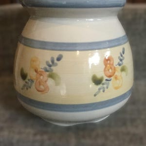 May include: A white ceramic vase with a blue rim and floral design. The vase has a rounded shape and a blue and yellow floral pattern.