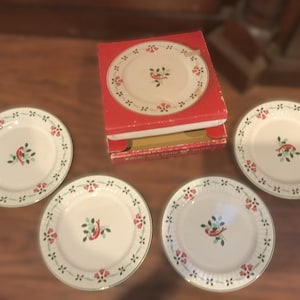 May include: Set of four white ceramic plates with a red bird and floral design. The plates are arranged around a red box containing additional plates with the same pattern. The plates have a gold trim.