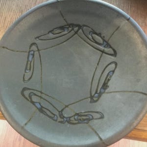 May include: A round, gray ceramic plate with a decorative design. The plate features a series of black, curved lines and shapes with blue accents. The plate is a neutral color and appears to be handmade.