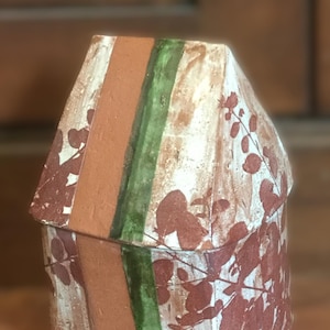 May include: A handmade ceramic house-shaped box. The box features a terracotta-colored central stripe, flanked by a green stripe and white panels with a reddish-brown leaf pattern. The lid mirrors the design. The box is on a wooden surface.