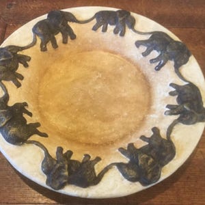 May include: A white ceramic dish with a raised rim featuring a circle of black elephant figurines around the edge. The dish has a light brown center.