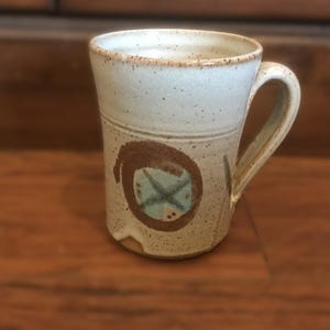 May include: A white ceramic mug with a brown and blue design. The mug has a handle and a speckled glaze.