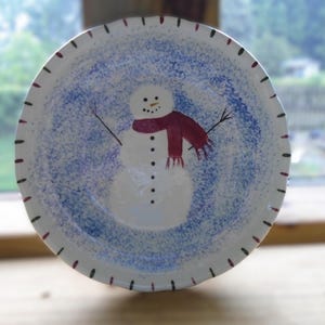 May include: A white ceramic plate with a hand-painted snowman design. The snowman has a red scarf, black coal eyes and buttons, and twig arms. The plate has a blue speckled background and a dotted rim.