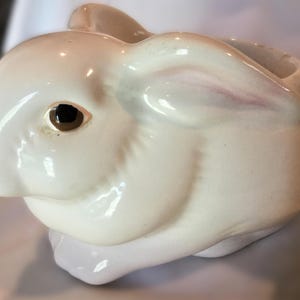 May include: A white ceramic bunny-shaped planter with a brown eye and pink accents on the nose and ears. The planter has a smooth, glossy finish and a hollow interior for holding plants or other items. The bunny is in a seated position.