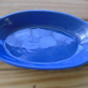 May include: A cobalt blue oval ceramic baking dish with two handles. The dish has a glossy finish and is empty. The dish is sitting on a wooden surface.