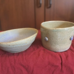 May include: A ceramic bowl and a matching ceramic cup with a speckled beige glaze. The bowl has a shallow, oval shape with a light interior, while the cup is cylindrical with small, raised blue decorative dots on the exterior surface.