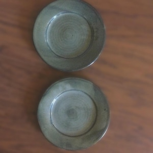 May include: Two round, ceramic plates with a muted green glaze. The plates have a slightly raised rim and a shallow center. The plates are displayed on a brown wooden surface.