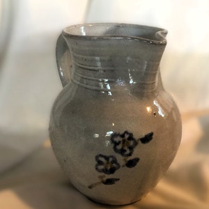 May include: A small, white ceramic pitcher with a floral design. The pitcher has a slightly flared lip and a handle that curves back towards the body. The flowers are painted in shades of blue and brown.