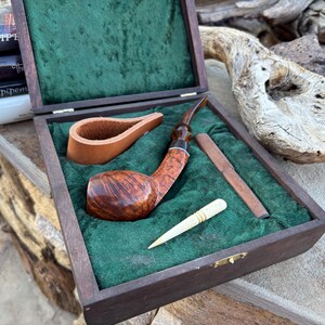 Pode incluir: Uma caixa de madeira com um forro de veludo verde contém um cachimbo marrom e preto, um socador de madeira e uma ferramenta de limpeza branca. A caixa é feita de madeira escura e tem uma alça de couro.