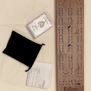 May include: A wooden cribbage board with a dark brown stain and the word "Kellner" engraved on the front. The board is accompanied by a deck of cards, a black velvet bag, and instructions for playing the game.