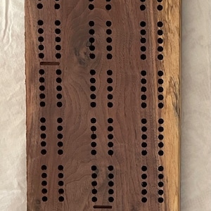 May include: A wooden cribbage board with a brown stain and a unique grain pattern. The board has a traditional design with holes for pegs and a track for scoring. The board is personalized with the names "Don & Linda".