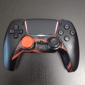 May include: A black and grey PlayStation 5 controller with a red and black patterned design. The controller has a black touchpad and a black button with a white PlayStation logo.